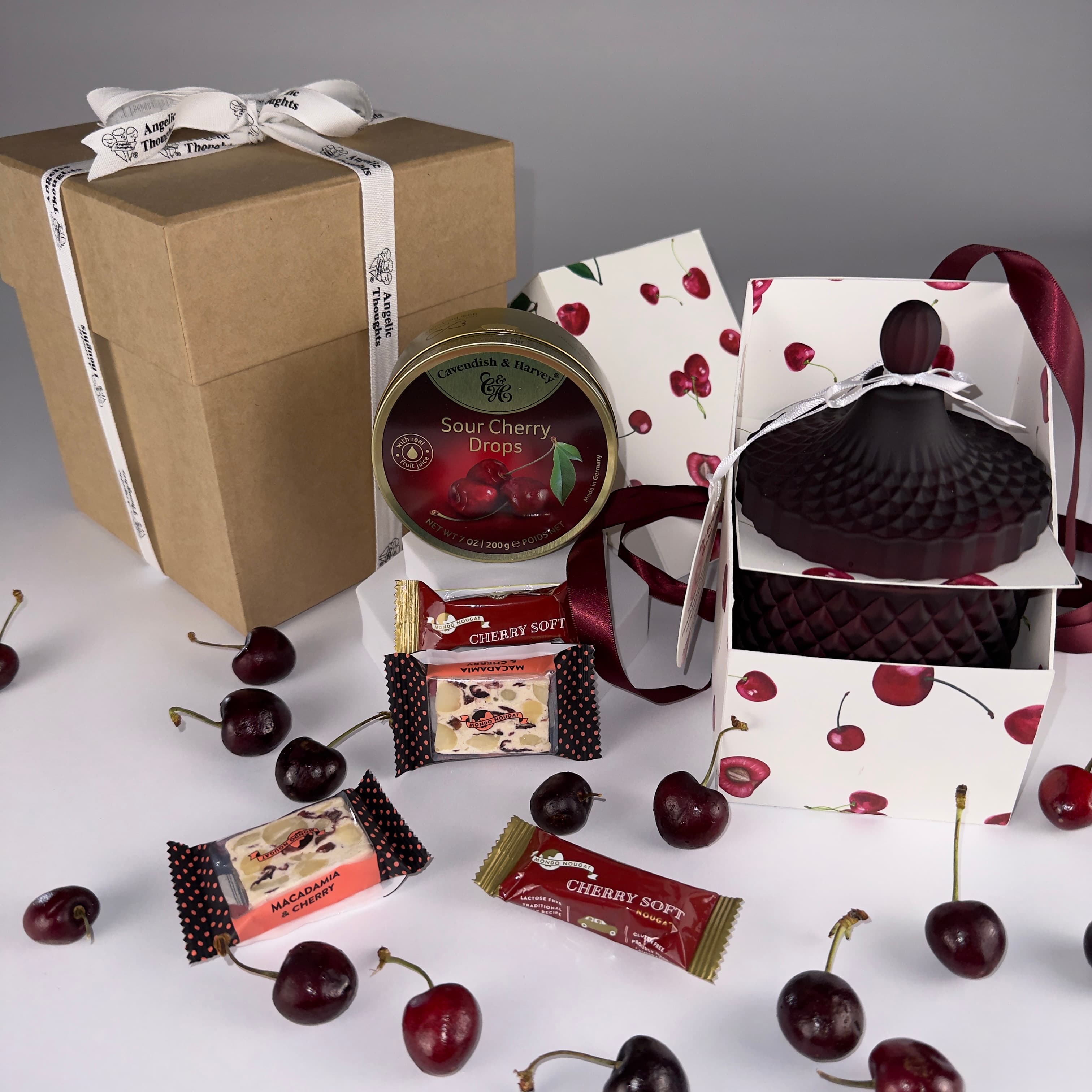 Angelic Thoughts Brown Kraft Gift Box Tied with Angelic Thoughts Ribbon,Cherish Love Cherry Love Gift Box Bundle Standard being displayed with loose cherries scattered amongst products which are: Sour Cherry Drops Tin, Cherry Soft Nougat, Cherry & Macadamia Nougat, Hand poured Soy Wax Cherry Candle in Art Deco Jar with personalised label
