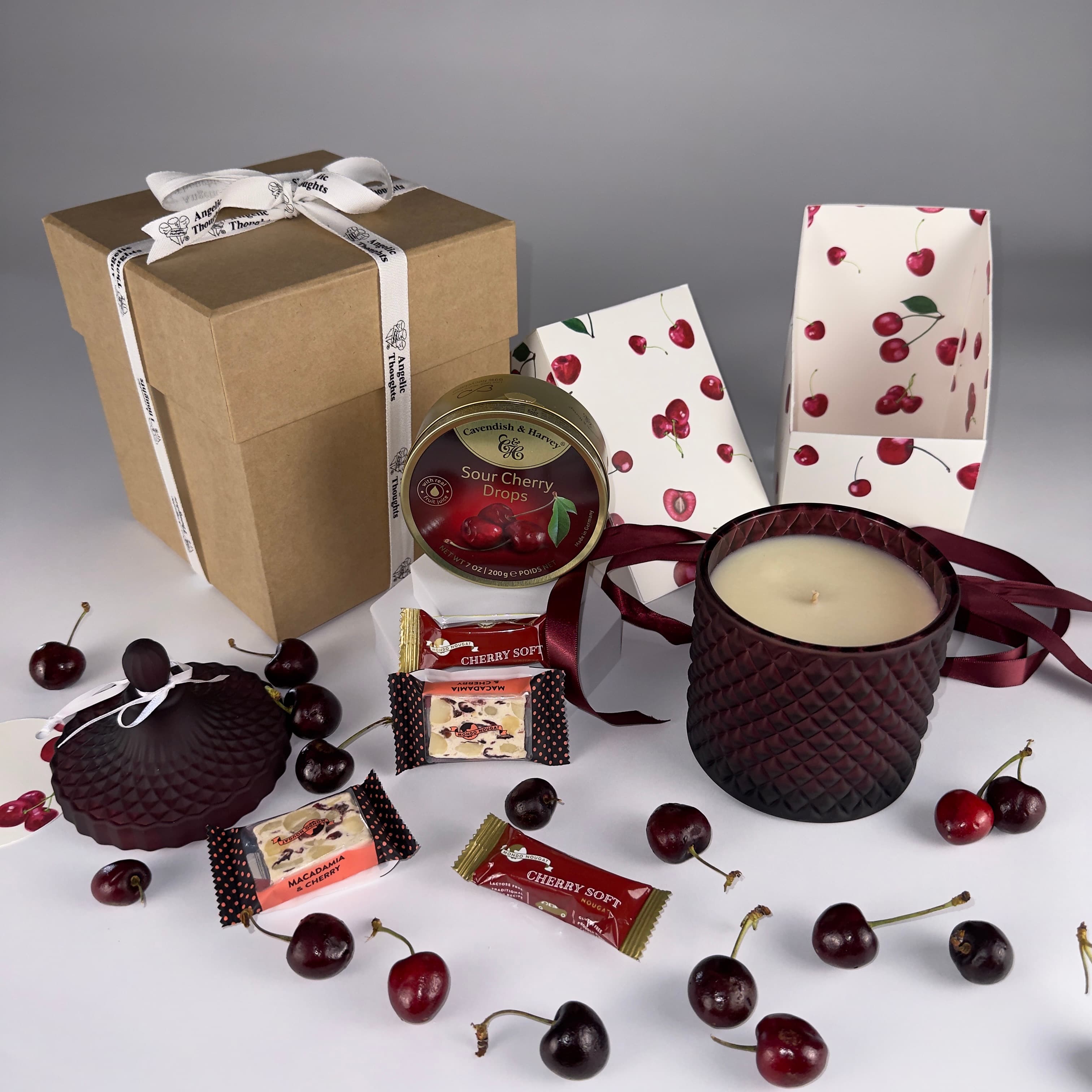 More open view of Angelic Thoughts Brown Kraft Gift Box Tied with Angelic Thoughts Ribbon,Cherish Love Cherry Love Gift Box Bundle Standard being displayed with loose cherries scattered amongst products which are: Sour Cherry Drops Tin, Cherry Soft Nougat, Cherry & Macadamia Nougat, Hand poured Soy Wax Cherry Candle in Art Deco Jar shown with lid off - lid has personalised label