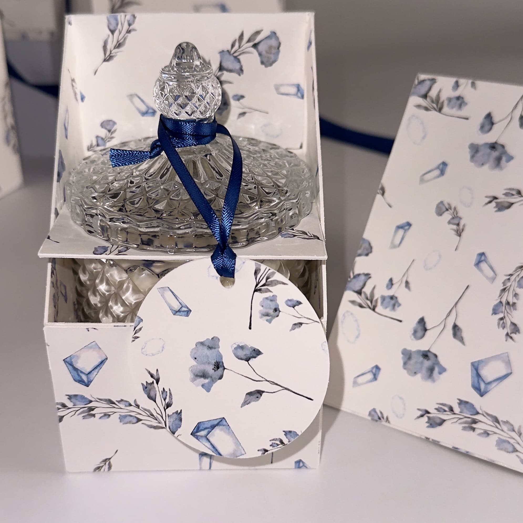 Angelic Whispers floral handpoured soy wax candle in Art Deco Crystal Glass embellished with lapus lazuli. The lid is on with a label tied in navy blue ribbons sitting beside the candle infront of an open product box with a rustal and floral print