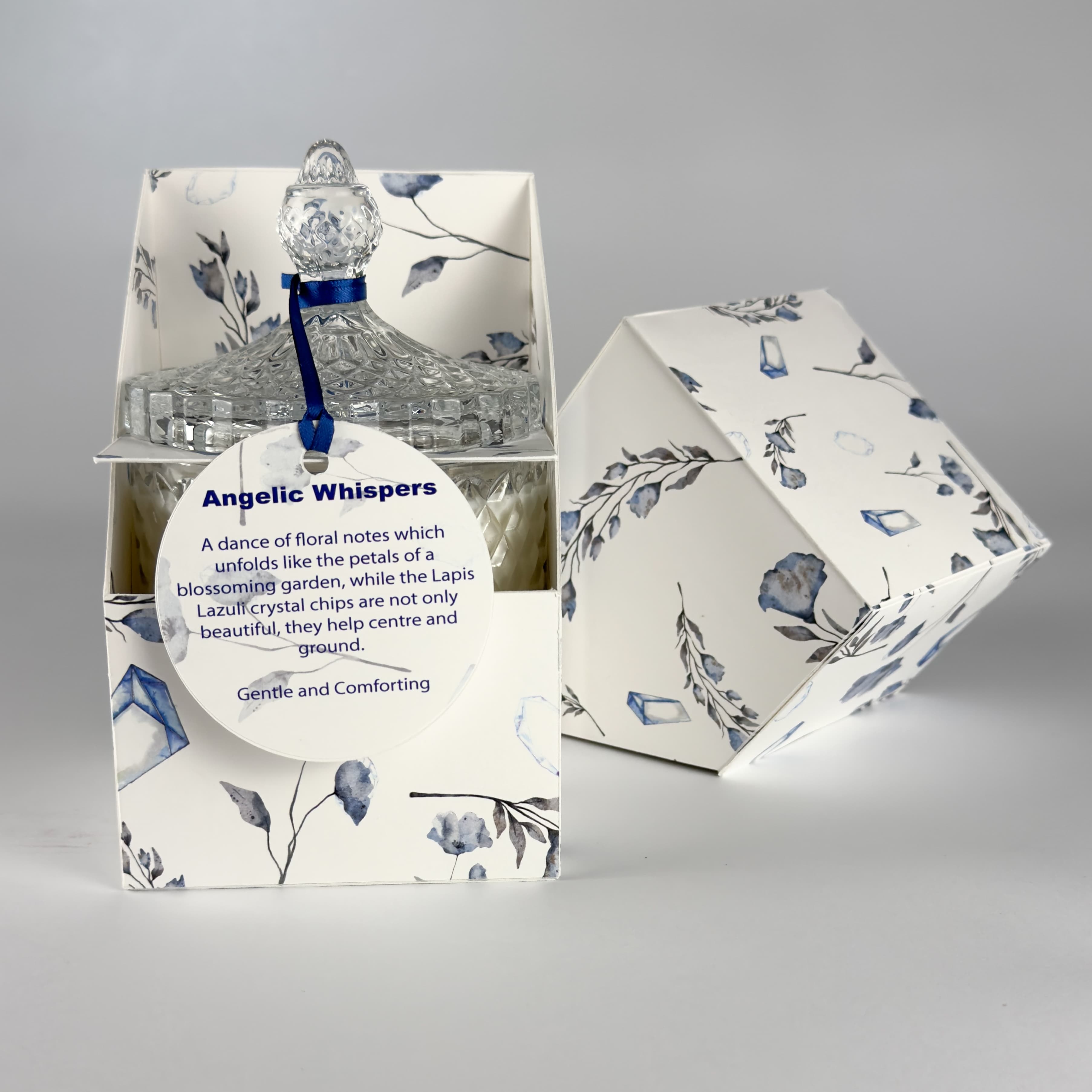 Angelic Whispers floral scented candle displayed inside an open, floral print gift box. The attached round label reads &