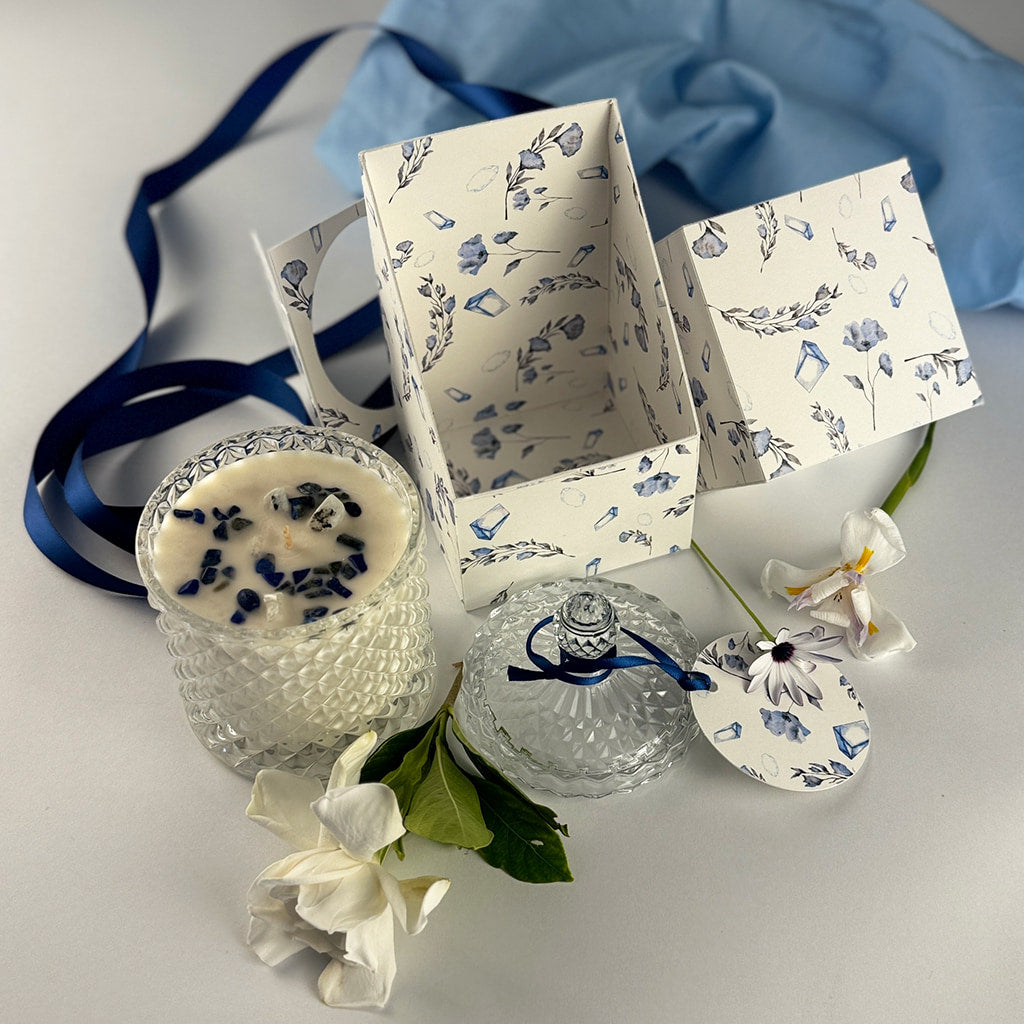 Floral handpoured soy wax candle in Art Deco Crystal Glass embellished with lapus lazuli. The lid is off and beside it with a label tied in navy blue ribbons sitting beside the candle infront of an open product box with a rustal and floral print and fresh flowers are scattered
