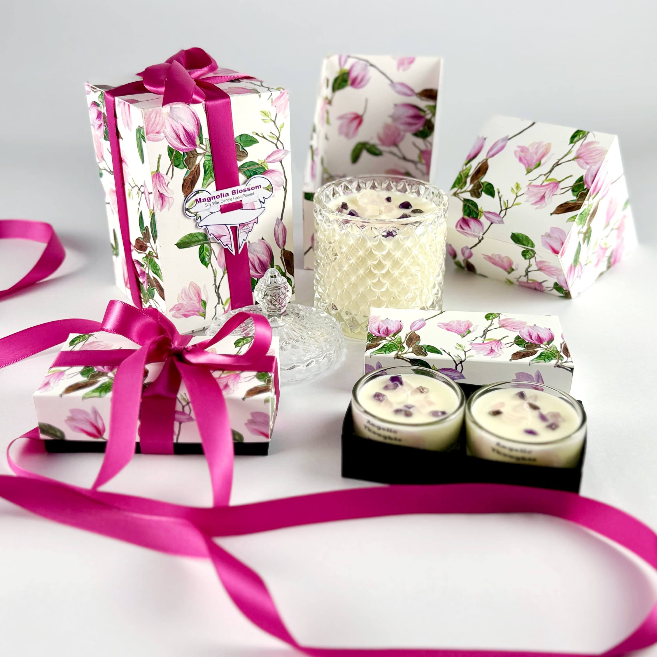 magnolia scented rose quartz and amethyst embellished hand poured tealights in glass tealight containers in black base box, with product box lid behind and with magnolia 2pack tealight product box and standard art deco Magnolia candle box both tied with deep pink ribbon on the left side. Lid off standard magnolia candle and open product box behind