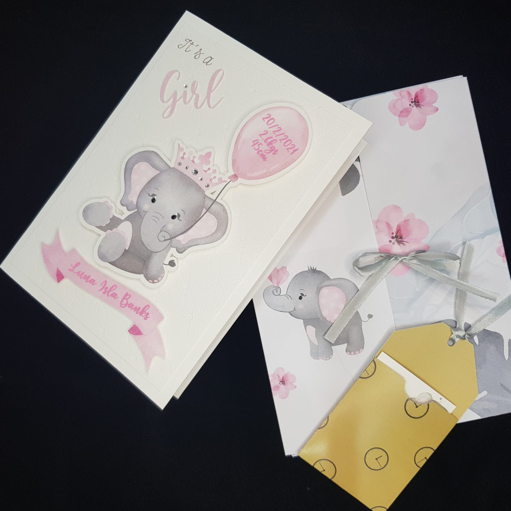 Image of the baby girl grey elephant card and a folder. The image of baby girl card is on the left. The Folder image has an image of a small grey elephant, scattered pink flowers and a grey ribbon tie to keep folder closed. There is also an image of the gift tag holder containg the white gift tag. The holder is a mustard colour.
