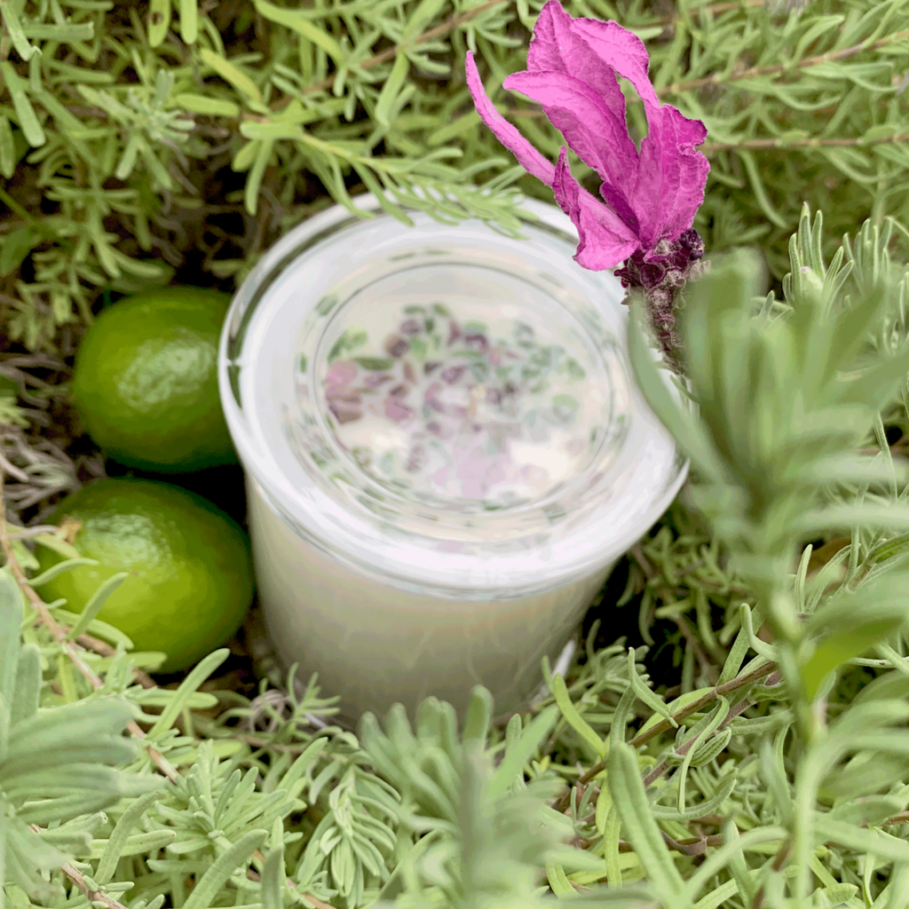 top down view of lavender and lime scented soy candle with lid on nestled in lavender bush with lavender flower - amethyst and green aventurine crystals embellish the candle and two limes site beside