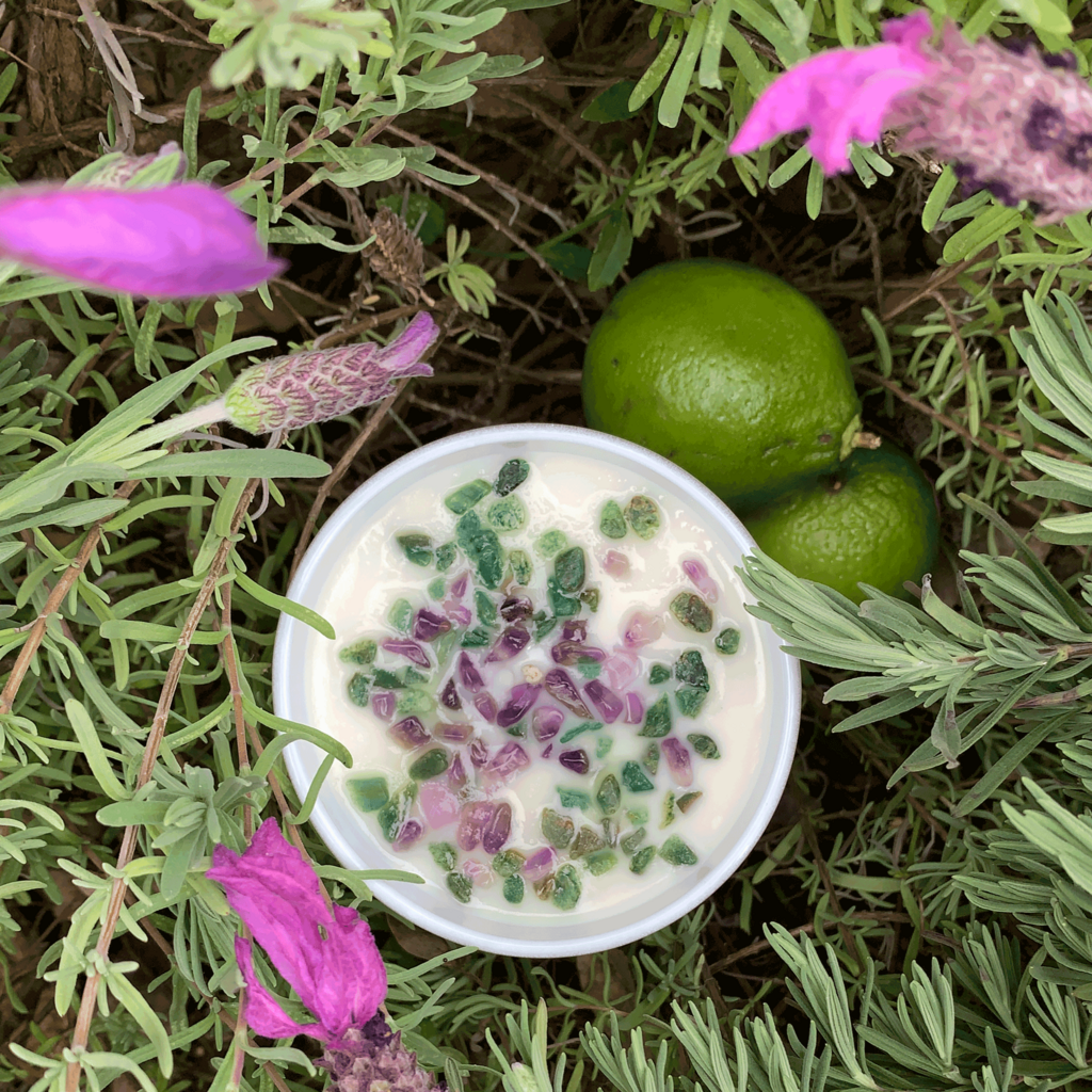 top down view of lavender and lime scented soy candle nestled in lavender bush with lavender flowers- amethyst and green aventurine crystals embellish the candle and two limes site beside