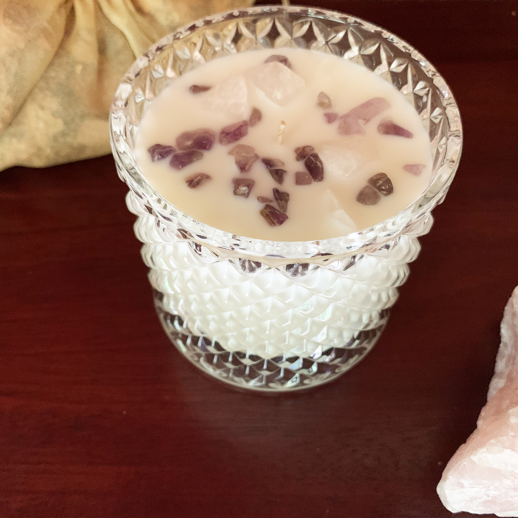 Elevated front view of Magnolia blossom candle in clear art deco style jar embellished with rose quartz and amethyst crystal chips. It is on a wooden table with a piece of rose quartz visible in the lower right