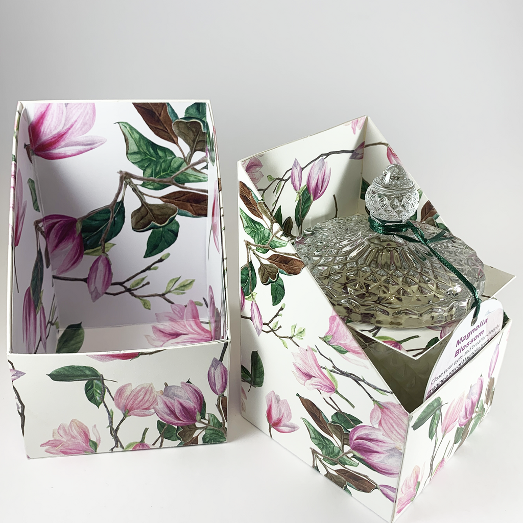 magnolia blossom product box lid to the left and box base to the right containing scented rose quartz and amethyst embellished hand poured soy wax candle with lid on in clear glass art deco style container. Magnolia Blossom product label is tucked into the front of the box base