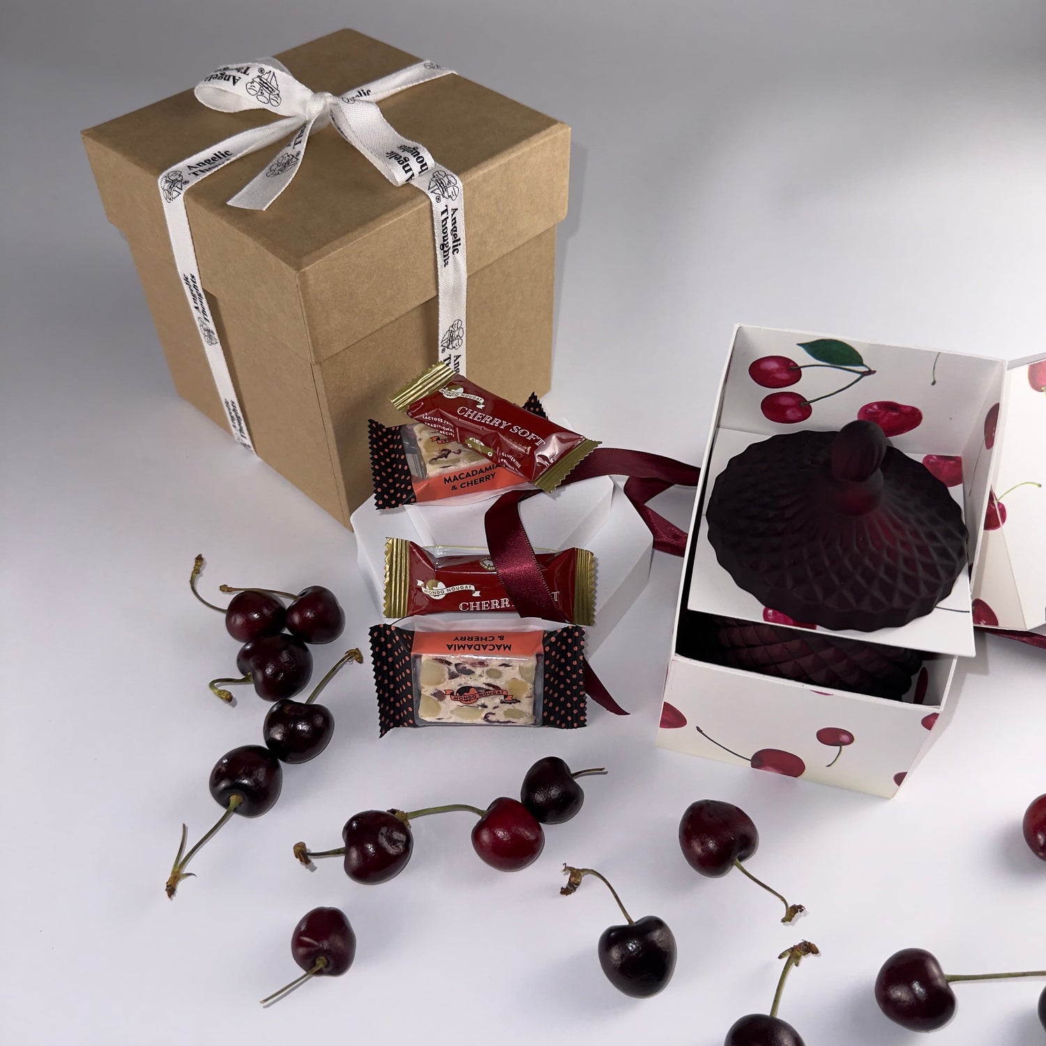 Angelic Thoughts Brown Kraft Gift Box Tied with Angelic Thoughts Ribbon,Cherish Love Cherry Love Gift Box Bundle Standard being displayed with loose cherries scattered amongst products which are: 2 x Cherry Soft Nougat, 2 x Cherry &amp; Macadamia Nougat, Hand poured Soy Wax Cherry Candle in Art Deco Jar with lid on
