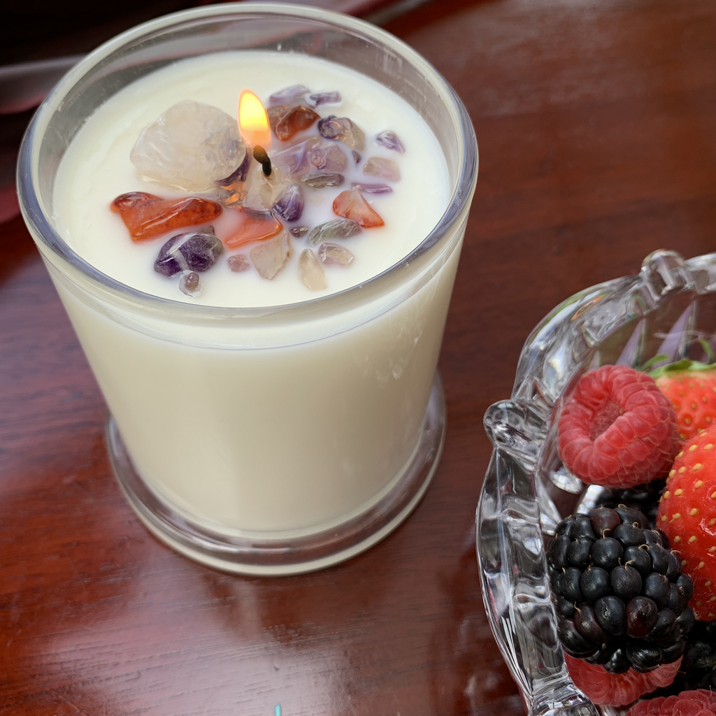 flickering flame on Berry Delightful Soy Wax candle in a clear glass container embellished with Clear Quartz, Amethyst and Carnelian Chips. Part of a crystal bowl containing raspberries, blackberries, strawberries and blueberries is visible in the bottom right corner