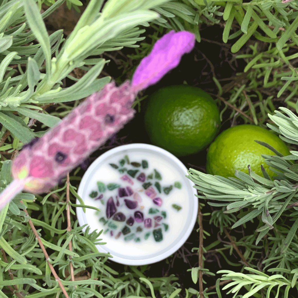 top down view of lavender and lime scented soy candle with lid on nestled in lavender bush with lavender flower - amethyst and green aventurine crystals embellish the candle and two limes site beside