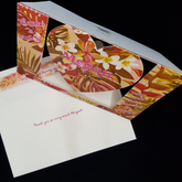 A beautiful heart swing card stands up on a matching envelope. with the words "thanks a bunch" on the left, the name Megan sits centrally on the swinging heart and another sentiment "your kind-ness means so much" sits on the right.  Tropical flowers and foliage are the focus of this muted green and pink toned background. Pops of pearl colours including yellow, orange and pinks have been selectively hand applied.
